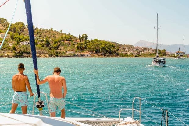 Photo of Greek Saronic Islands sailing with two males standing on a MedSailors catamaran approaching Poros Greece. 